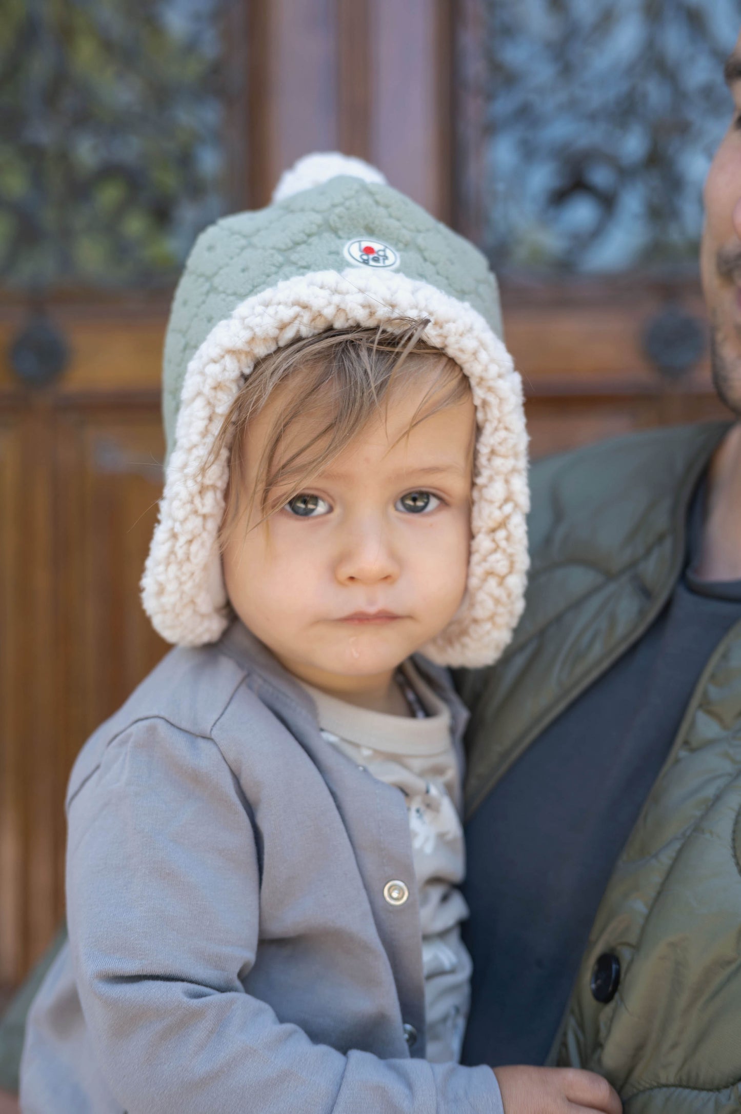 Lodger Hatter Folklore Fleece cepure drizzle krāsa, bildē redzams mazulis ar cepuri galvā un pieagušā rokās.